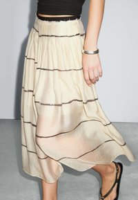 Jupe midi plissée crème avec des rayures horizontales marron, fabriquée en tissu léger. Présente une silhouette décontractée et une texture douce.