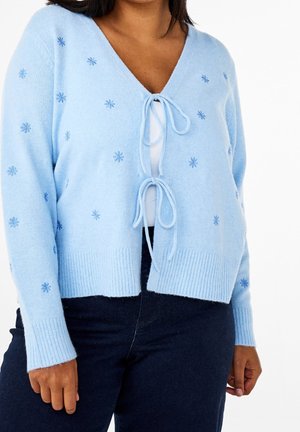 Frau trägt hellblauen gestrickten Cardigan mit kleinen bestickten Schneeflocken und zwei vorderen Bändern über einem weißen Top und dunklen Hosen.