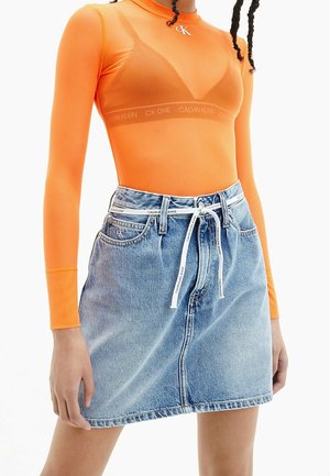 Kobieta w pomarańczowym prześwitującym topie z długim rękawem na biustonoszu Calvin Klein, w połączeniu z jasnoniebieską dżinsową spódnicą i paskiem marki Calvin Klein.
