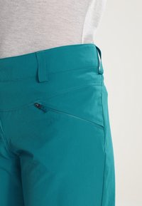 Turkosbyxor med en slät yta, utrustade med en framficka med blixtlås, bältesögon och skräddarsydda sömmar längs midjebandet.