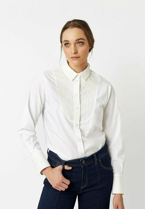 Eine Frau mit hellbraunen, nach hinten gebundenen Haaren trägt ein weißes, strukturiertes Hemd mit Knöpfen und dunkle blaue Jeans. Sie steht vor einem schlichten Hintergrund.