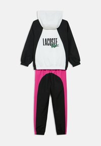 Jachetă alb-negru cu textul „LACOSTE” și logo verde, asortată cu pantaloni negri cu o curbă roz mare pe lateral.