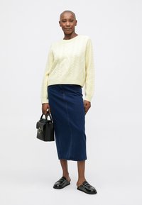 Nainen, joka on pukeutunut kermaväriseen palmikkoneuleeseen ja siniseen midi-hameeseen, pitää mustaa käsilaukkua kädessään. Hänen jalkojaan verhoavat mustat slip-on-loaferit, ja hän seisoo valkoista taustaa vasten.