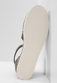 Semelle d’une sandale noire avec un bord tressé marron style espadrille et une semelle blanche en caoutchouc striée, tournée vers l’avant.