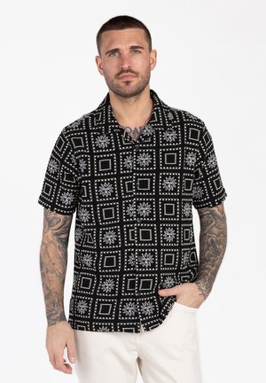 Mann mit kurzen Haaren und Tattoos, trägt ein schwarzes kurzärmeliges Hemd mit Muster und weiße Hose, steht mit einer Hand in der Tasche.