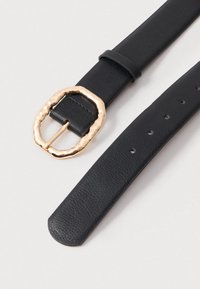 Melna ādas jostā ar teksturētu apdari, liela, nevienmērīgi veidota zelta sprādze un vairāki regulēšanas caurumi.