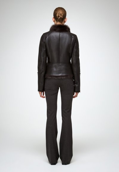 Donna in piedi di spalle, che indossa una giacca di pelle marrone scuro con rifiniture in pelliccia e pantaloni grigi scuri a zampa d'elefante, su uno sfondo bianco semplice.