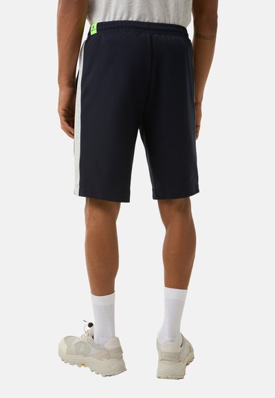 Marineblau Shorts mit weißen Seitenstreifen, elastischem Bund und grünem Akzentetikett. Getragen mit weißen Socken und hellen Turnschuhen.