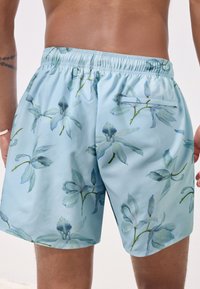 Șort de baie floral albastru cu textură moale, având un model cu flori verde deschis și albastre, talie elastică și un singur buzunar în spate.