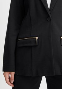 Černý blazer s přizpůsobeným střihem a jedním knoflíkem, doplněný o dvě zipové kapsy s pozlacenými detaily. Měkká tkanina.