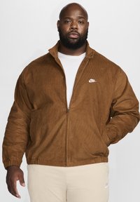 Brūns kordūras džemperis ar rāvējslēdzēju un augstu apkakli, ar elastīgām aprocēm. Uz kreisā krūšu paša redzams balts Nike logo. Valkāts virs baltas krekla.