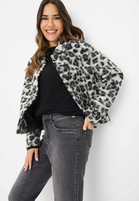 Donna sorridente che indossa un top nero, jeans grigi e un giacca pelosa con stampa animale bianca e nera, mentre posa con una mano in tasca.