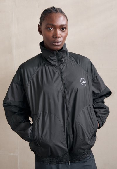 Veste coupe-vent noire en nylon avec un col haut, des poignets élastiques et un logo du côté gauche. Texture lisse et coupe décontractée.