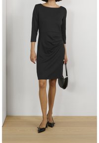 Robe noire avec un col bateau, des manches trois-quarts, des détails froncés sur le côté et une coupe ajustée, associée à un sac à main noir et des talons.