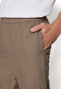Hnedé nohavice s elastickým pásom a hladkou textúrou, so bočnými vreckami. Módny strih s jemným leskom. Nosí sa s jednoduchým bielym tričkom.