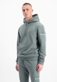 Lichtgroene hoodie van zacht materiaal, met een voorzak, verstelbare capuchon en een witte tekstlogo op de rechtermouw.