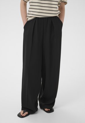 Personne portant un pantalon noir ample, une chemise beige à rayures, des sandales noires et se tenant debout, les mains dans les poches, devant un fond uni.