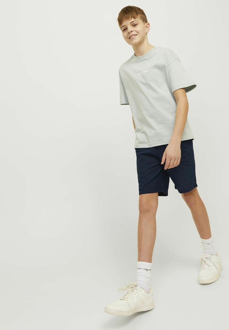 T-shirt en coton gris clair avec un col rond et des manches courtes, associé à un short bleu marine et des baskets blanches, avec une marque discrète sur le t-shirt.