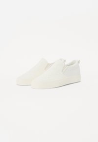 Hvite slip-on sko med teksturert blondermønster og solide paneler. Myk tekstiloverdel, gummisåle og minimalistisk design.