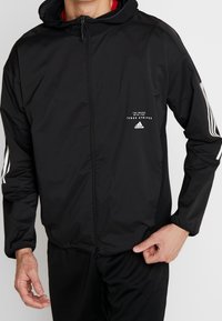 Svart zip-up jacka gjord av lättviktsmaterial, har en huva, vita randdetaljer på ärmarna och en liten logotyp på bröstet.