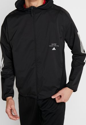 Zwarte zip-up jas gemaakt van lichtgewicht stof, heeft een capuchon, witte streepaccenten op de mouwen en een klein logo op de borst.