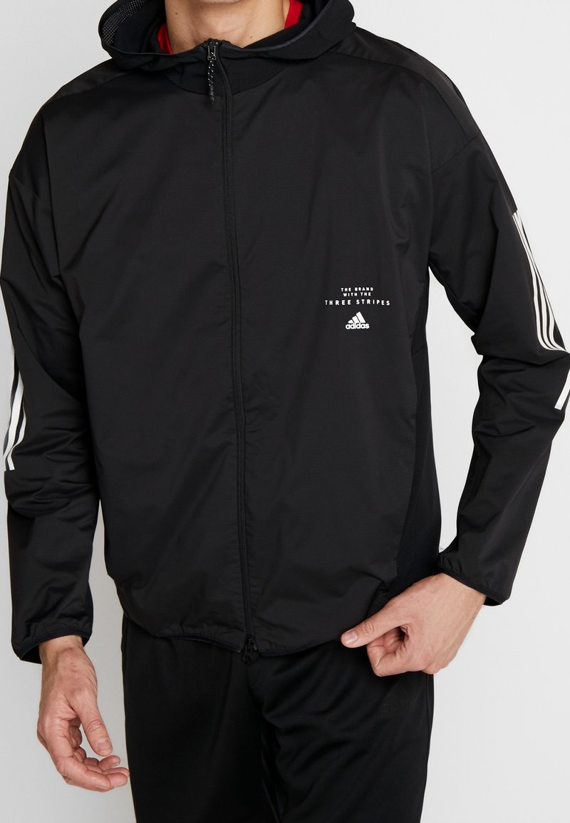 Svart zip-up jacka gjord av lättviktsmaterial, har en huva, vita randdetaljer på ärmarna och en liten logotyp på bröstet.