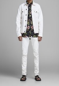 Giacca di jeans bianca sopra una camicia nera a maniche corte con fiori, abbinata a pantaloni bianchi slim-fit e sandali neri. Texture liscia, motivi vivaci.