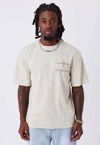 T-shirt beige a maniche corte con tessuto testurizzato, caratterizzato da un design del logo discreto sul petto, abbinato a jeans di denim azzurri chiari.
