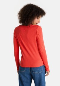 Eine Frau mit lockigem Haar, die ein langärmliges rotes Shirt und blaue Jeans trägt, steht mit dem Rücken zu einem schlichten weißen Hintergrund.