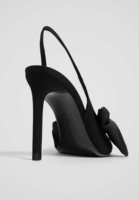 Zwarte slingback pump met hoge hak en satijnen afwerking, voorzien van een strikaccent aan de voorkant en een stilettohak. Soepel, elegant ontwerp.