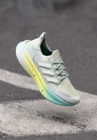 Light beige running shoe with three side stripes and gradient yellow to blue sole, shown in mid-air above textured asphalt surface.