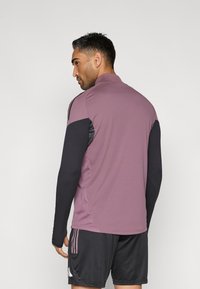 Αθλητική μπλούζα με ψηλό γιακά, με μωβ σώμα και μαύρα μανίκια. Διαθέτει 3 ρίγες στον ώμο και υφή υφάσματος.