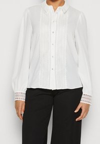 Blouse blanche à manches longues, fermeture boutonnée à l'avant, détails en dentelle au centre et aux poignets des manches, tissu doux, design à col.