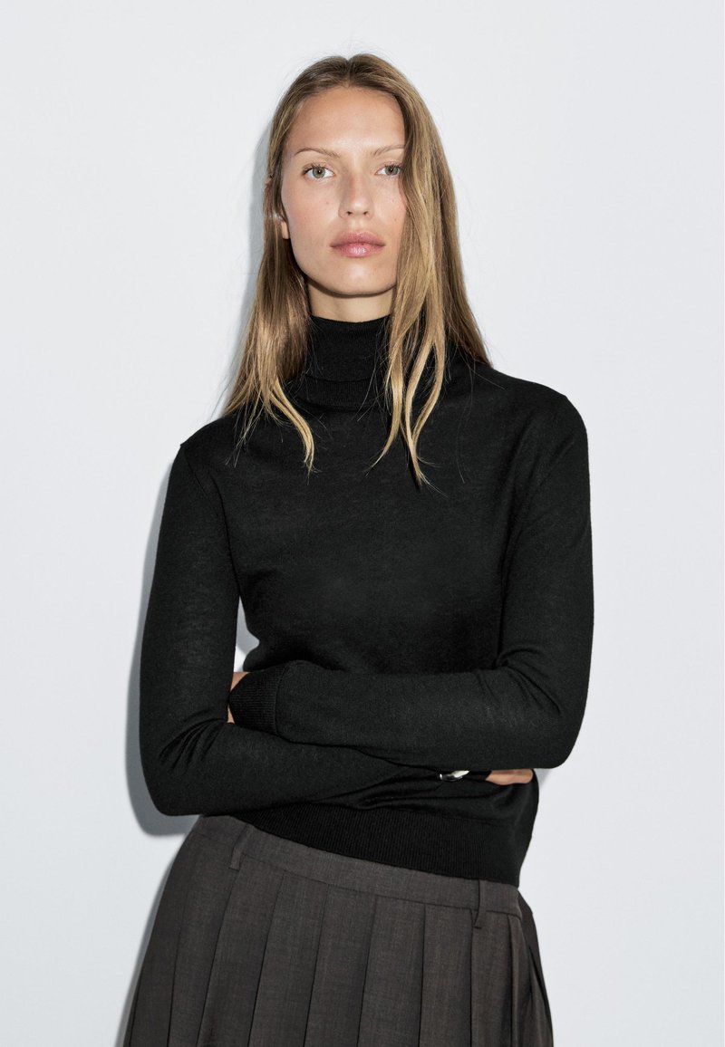 Maglione nero a collo alto con maniche lunghe, polsini e orlo a costine, abbinato a una gonna grigia plissettata. Sfondo neutro.