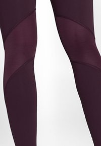 Legginsy z ciemnofioletowej tkaniny i jaśniejszych fioletowych paneli siatkowych tworzących geometryczne wzory na dolnej części nóg.