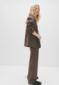 Žena sa dugom plavom kosom koja nosi labavi smeđi odijelo i crne cipele, stoji u minimalnom bijelom prostoru.