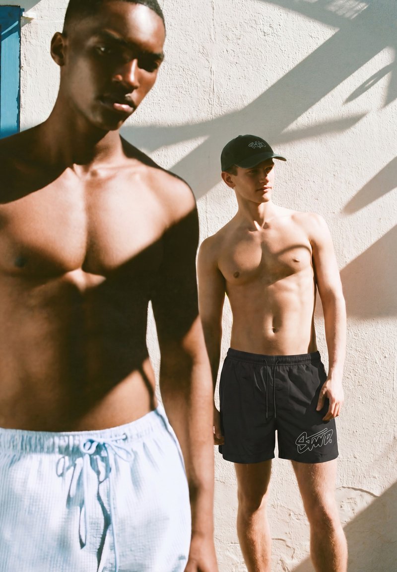 Deux hommes torse nu se tiennent contre un mur texturé ensoleillé, l'un portant un short bleu clair et l'autre un short noir et une casquette noire.
