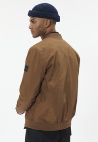 Chaqueta bomber marrón hecha de tela suave, con cuello y puños acanalados, y un parche negro en la manga izquierda.