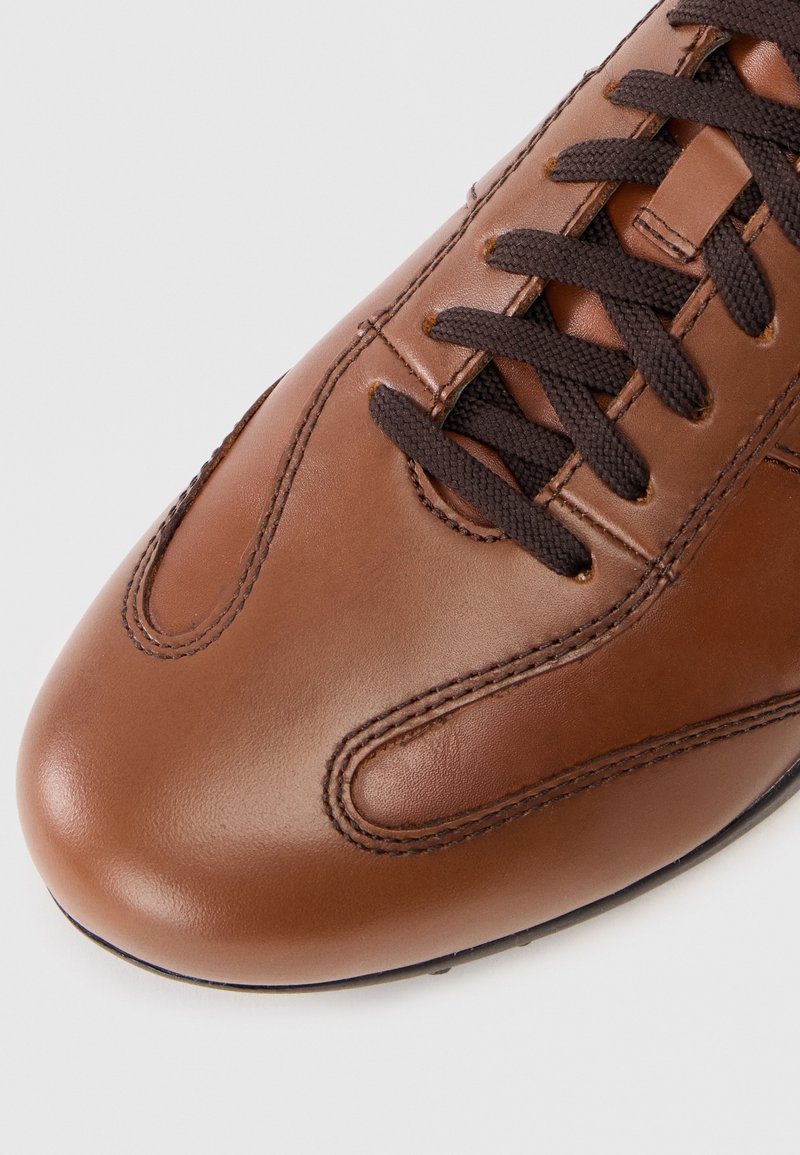 Brązowe skórzane sneakersy z ciemnobrązowym sznurowadłem, zaokrąglonym noskiem i subtelnymi detalami przeszycia. Gładka tekstura i elegancki design.