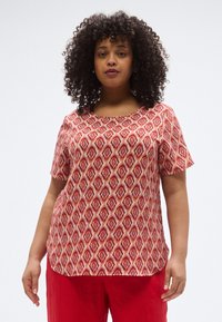 Κόκκινη κοντομάνικη μπλούζα με γεωμετρικό σχέδιο και κρεμ φόντο. Το ύφασμα φαίνεται ελαφρύ και υφασμένο.