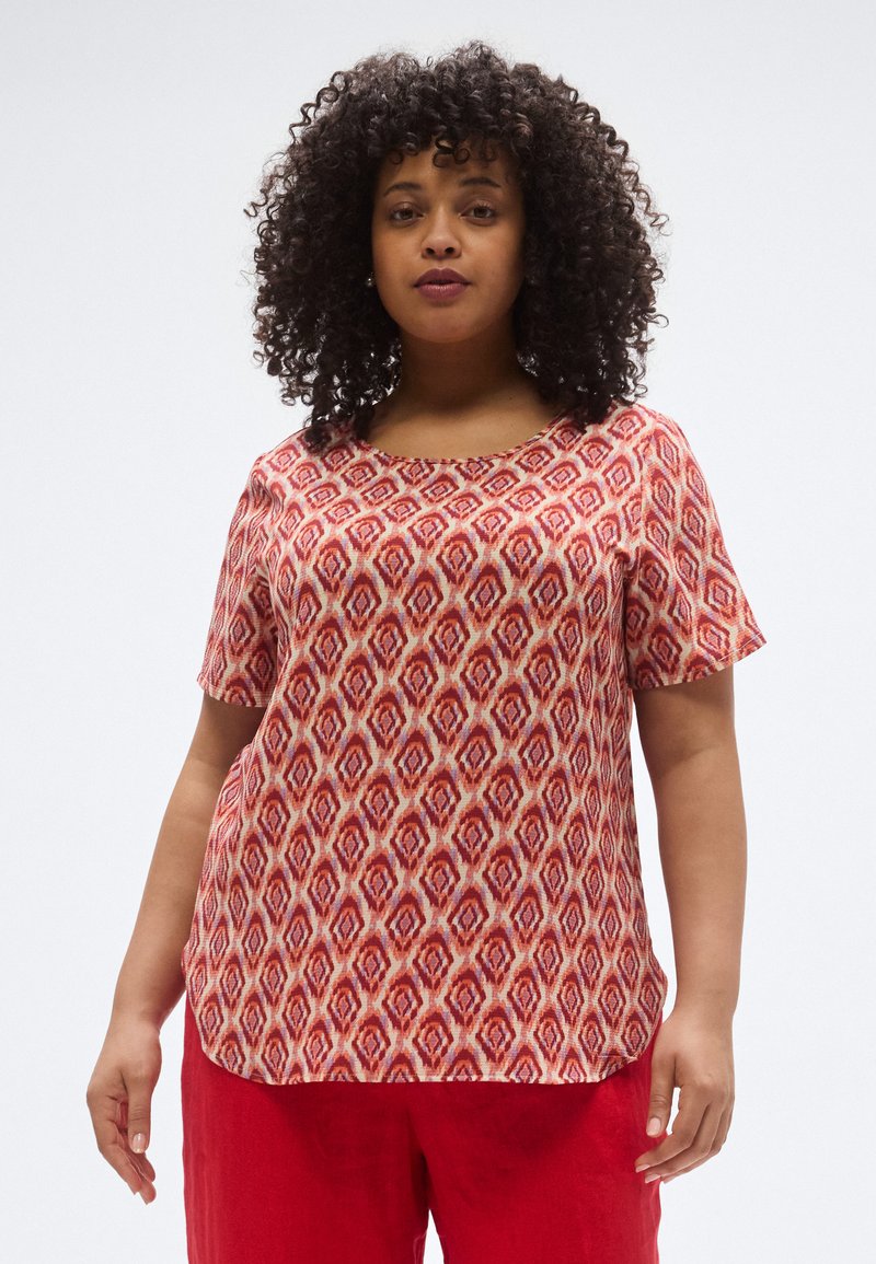 Κόκκινη κοντομάνικη μπλούζα με γεωμετρικό σχέδιο και κρεμ φόντο. Το ύφασμα φαίνεται ελαφρύ και υφασμένο.