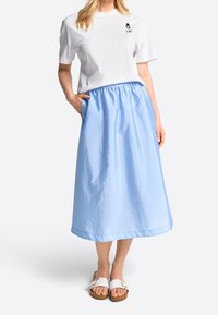 Mujer vistiendo una camiseta blanca con un pequeño gráfico y una falda midi azul claro brillante, de pie con una mano en el bolsillo de la falda y sandalias blancas.
