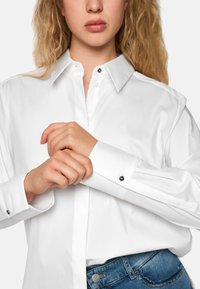 White long-sleeve shirt with a collar, featuring black button accents and cuffs. Paired with blue jeans, showing a smooth, sleek texture.