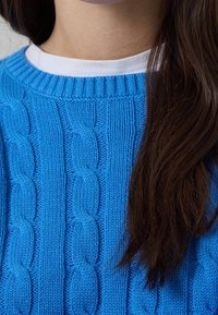 Helles blauer Kabelstrickpullover mit geripptem Ausschnitt, über einem weißen T-Shirt getragen. Die Struktur zeigt ausgeprägte Kabelmuster.