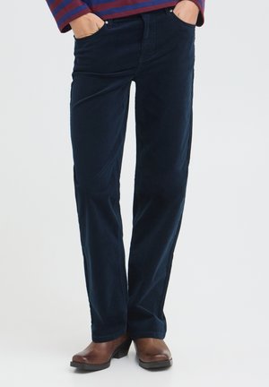 Personne portant un pantalon droit bleu foncé, des chaussures en cuir marron et une chemise rayée à manches longues, les mains dans les poches.