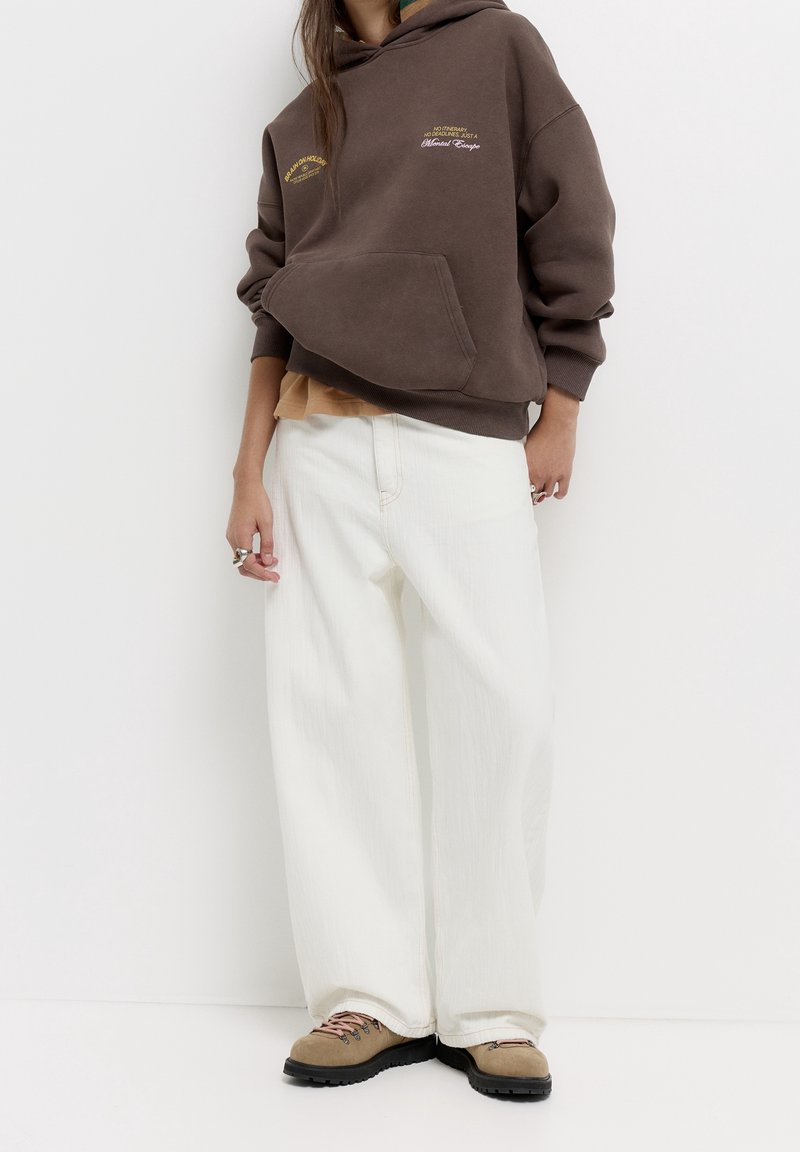 Personne portant un sweat à capuche marron, un pantalon blanc à jambes larges, des chaussures de randonnée beiges et des bagues à la main gauche sur un fond blanc.