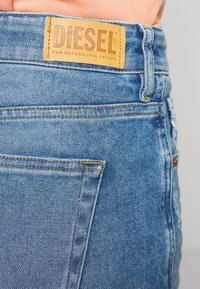Vaqueros de denim con un lavado azul claro, que presentan un parche de cuero marrón etiquetado como "DIESEL" y costuras amarillas en el bolsillo trasero.