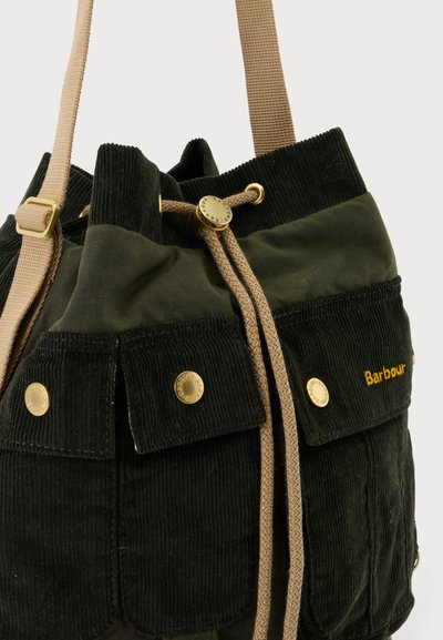 Кадър отблизо на тъмнозелена и черна кадифена торба Barbour с връзка за стягане, светлокафяви презрамки, обков в златист цвят и предни джобове с капаци.