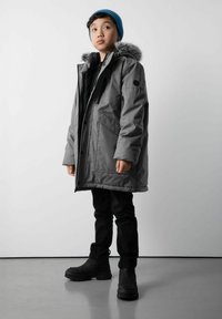 Młoda osoba ubrana w szary zimowy płaszcz z futrzanym kapturem, niebieską czapkę dzianinową, czarne spodnie i czarne buty, stojąca na szarej podłodze na tle białej ściany.