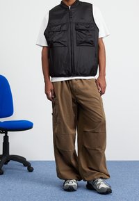 Gilet di nylon nero con due tasche frontali, indossato sopra una maglietta bianca. Pantaloni cargo marroni larghi e sneakers grigie completano l'outfit. Sedia blu nelle vicinanze.